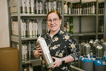 Lächelnde Frau hält eine kleine Gasflasche in der Hand, im Hintergrund Regel mit weiteren Gasflaschen