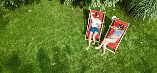 Zwei Frauen sitzen mit Drinks auf Liegestühlen im Garten auf grünem Rasen 
