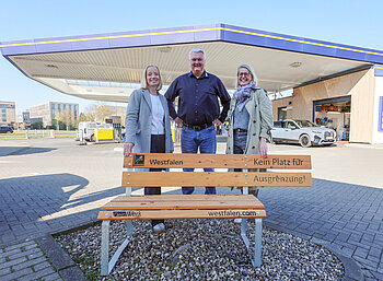 Zwei Frauen und ein Mann stehen hinter einer Bank an einer Tankstelle