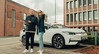 Ein Kollege und Kollegin stehen auf E-Auto Lade-Parkplatz vor dem weißen E-Auto Firmenwagen